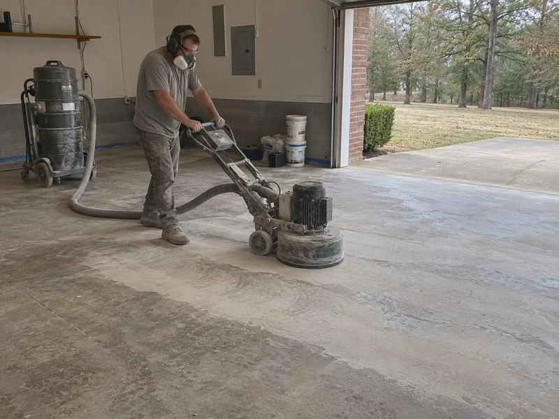 Worker in a respirator grinding a bare concrete garage floor with a dust vacuum behind